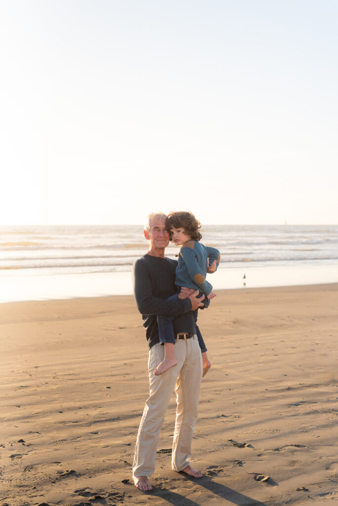 Grandpa holds grandson on the beach in later afternoon sun in Oceanside, CA