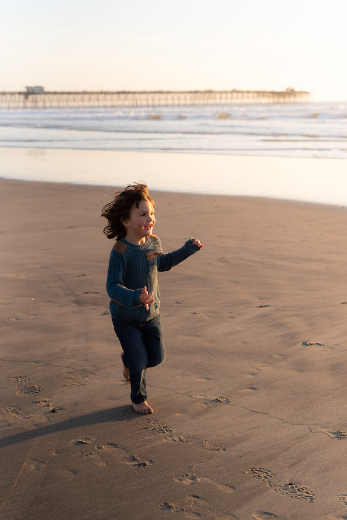 Young boy running down the beach and laughing with Oceanside Pier in background