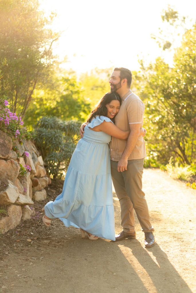 A couple bathed in golden light walk down a gravel path and the wife hugs the husband tightly while smiling at Alta Vista Botanical Garden