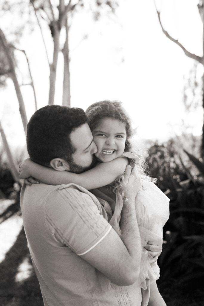 A father holds and kisses his young daughter in a black and white image while the little girl smiles at the camera at Alta Vista Botanical Garden