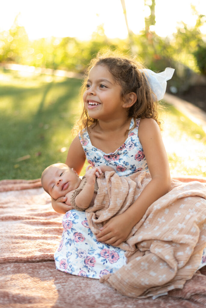 A little girl holds her newborn baby brother and smiles at someone off camera at Alta Vista Botanical Garden