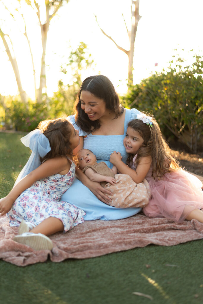 A mother sits with her two little girls and newborn son and smiles at one of the girls with soft golden light at Alta Vista Botanical Garden