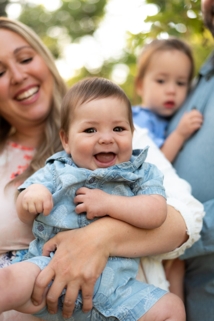 A candid moment of a happy baby smiling while his mother looks on and smiles behind him