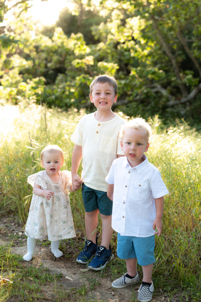 Three sibling holds hands on a path through spring green grasses at Marian Bear Park, San Diego