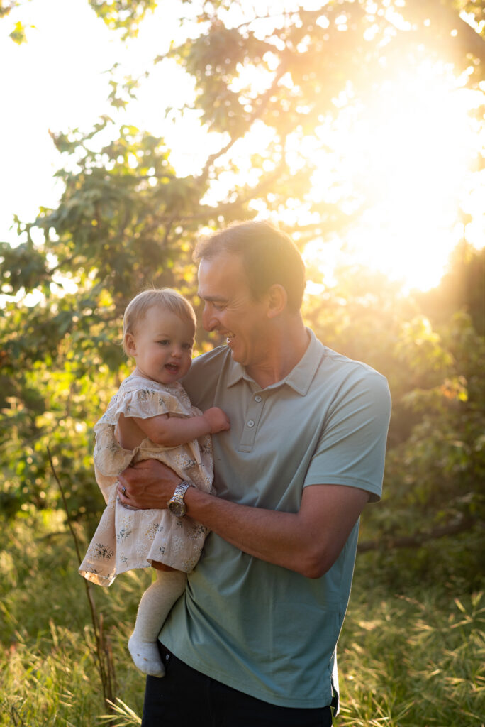 Dad holds his baby daughter while looking at and smiling at her with golden light filtering through the sycamore trees behind them at Marian Bear Park, San Diego