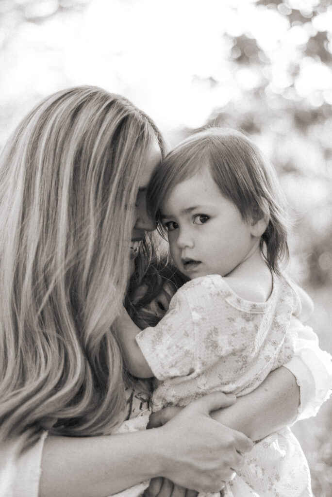Soft black and white image of a mother snuggling her toddler daughter while her daughter looks intently at the camera at Marian Bear Park, San Diego