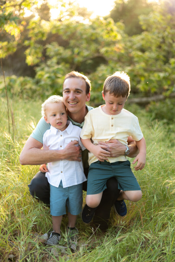 Dad crouches down to pose with his two sons in a green field with golden light filtering behind at Marian Beach Park, San Diego