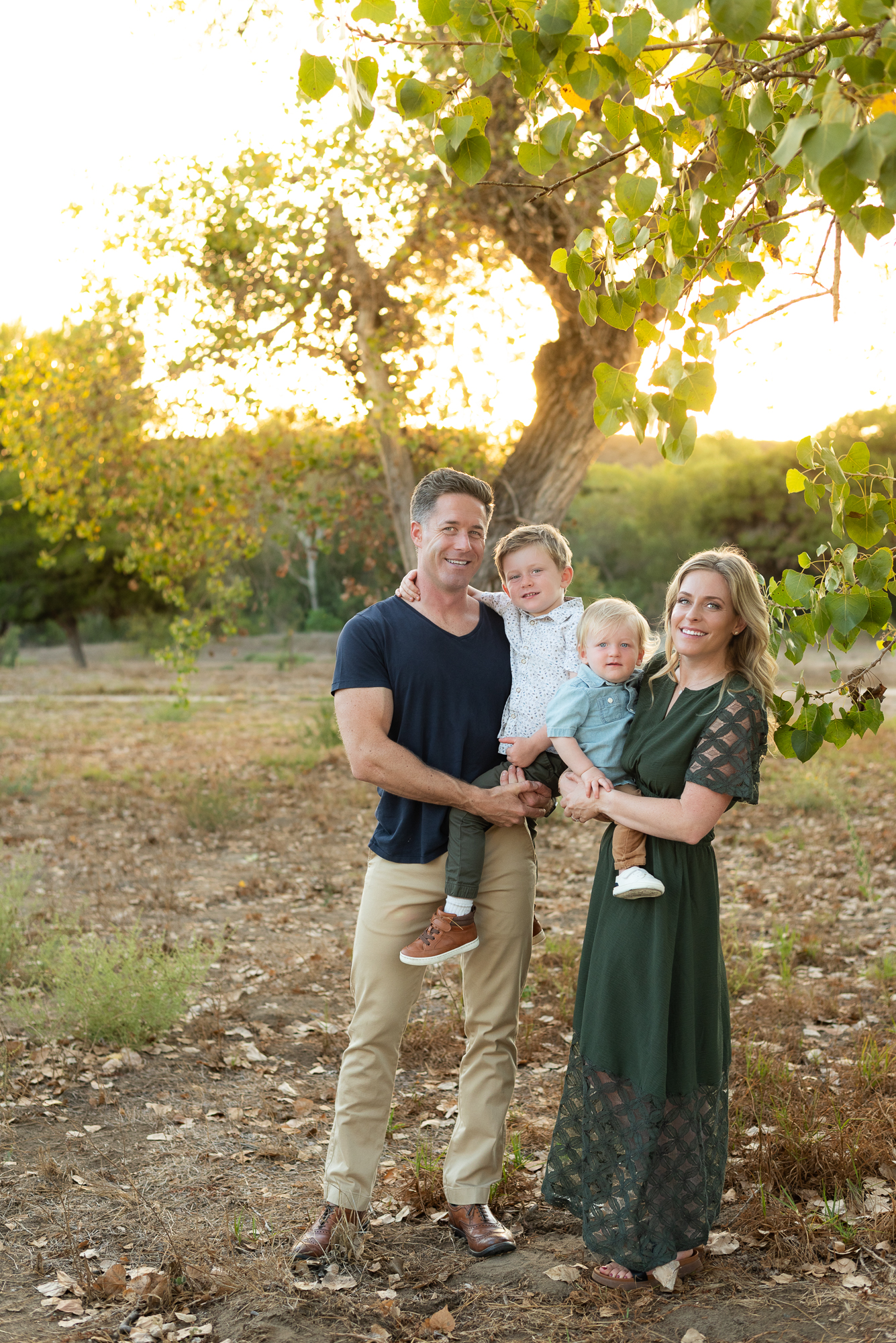 Family of four stands in a sycamore grove during golden hour with two young sons