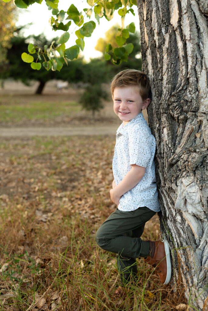 A 3 year old boy poses leaning against a tree with one foot up