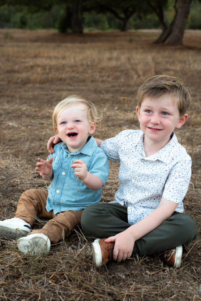 Two young brother sitting in a field laughing with trees behind
