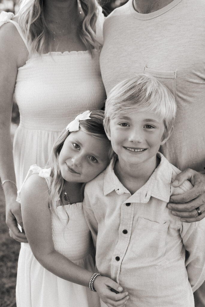 A black and white photo of a brother and sister hugging with their parents loving hands on their shoulders during their San Diego golden hour family photography session at Guajome Park, Oceanside