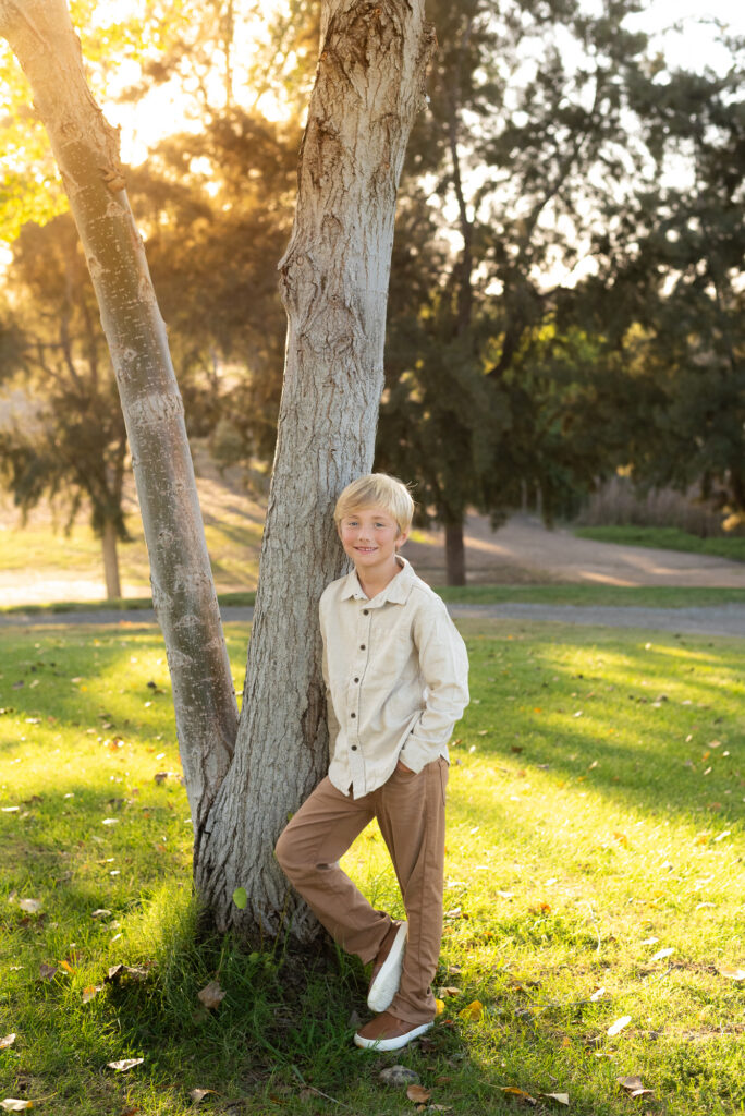 A little boy leans against a tree and smiles at the camera during their San Diego golden hour family photography session at Guajome Park, Oceanside