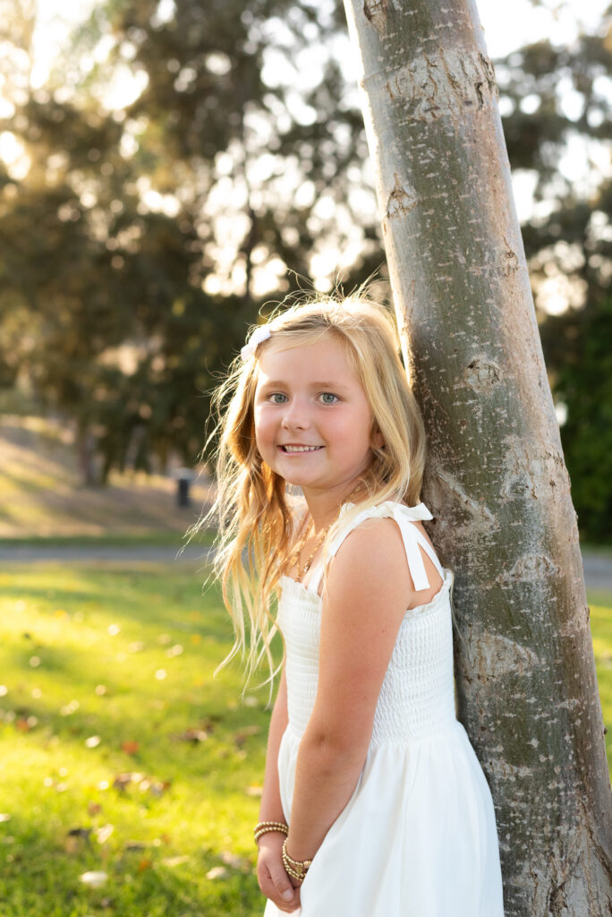 A little girl leans against a tree and smiles for the camera during their San Diego golden hour family photography session at Guajome Park, Oceanside