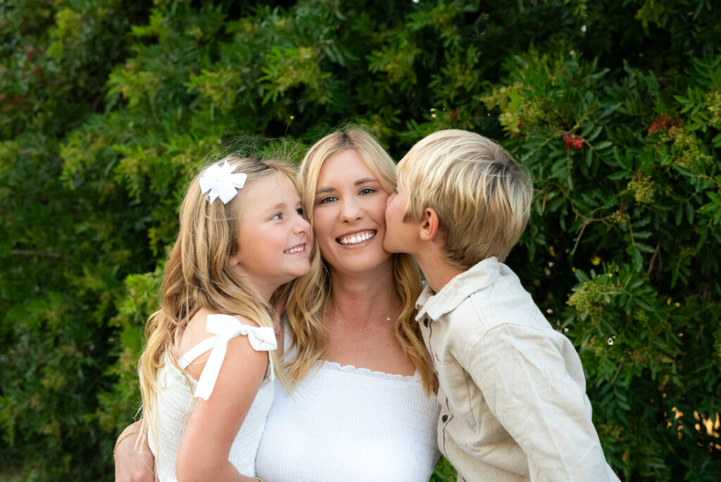 A boy and a girl kiss their mom on each cheek while she smiles at the camera during their San Diego golden hour family photography session at Guajome Park, Oceanside