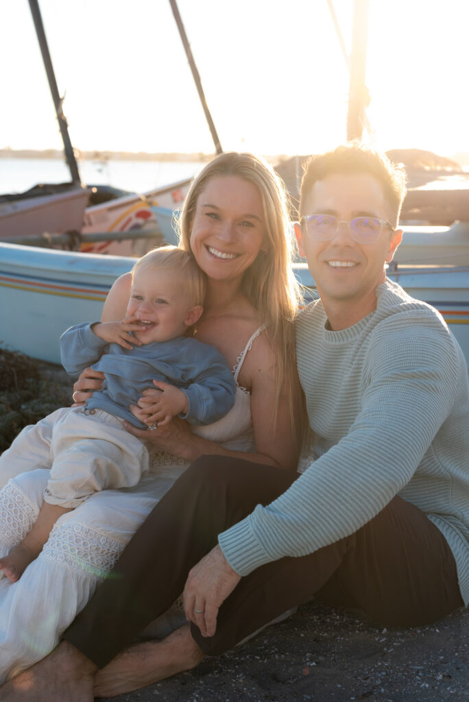 Family of three sit against a blue sailboat with their toddler son during golden hour in Pacific Beach, CA