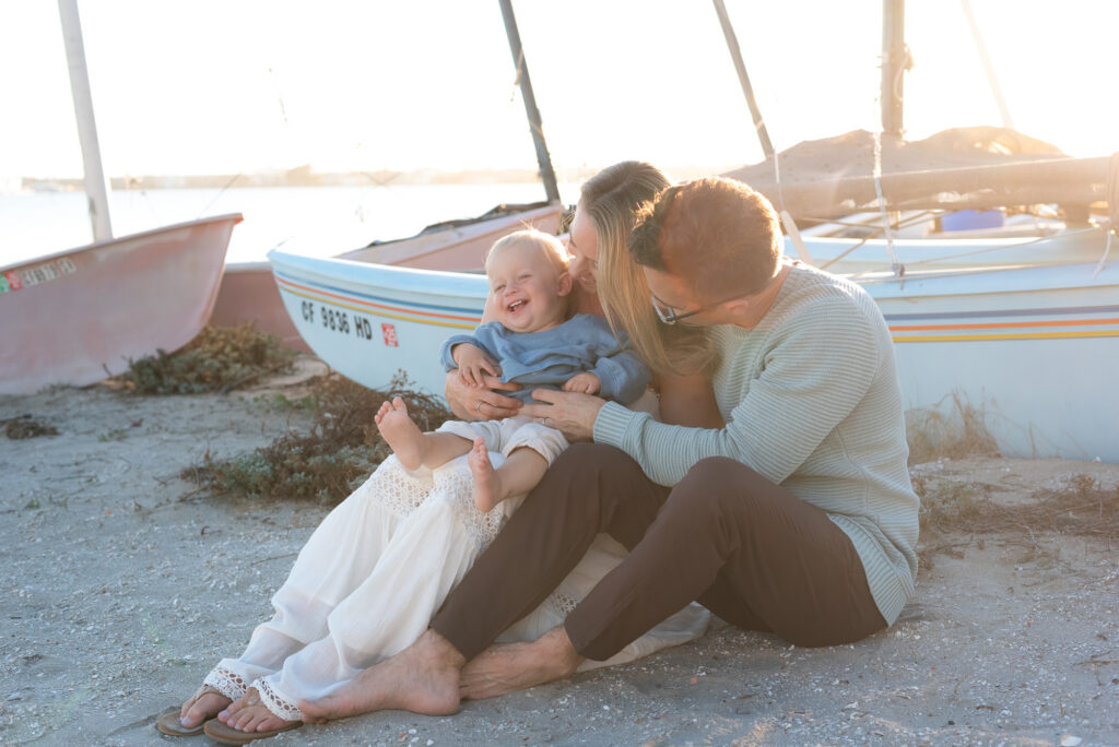 Family of three sits against a blue sailboat during golden hour at the beach with their toddler son during golden hour in Pacific Beach, CA