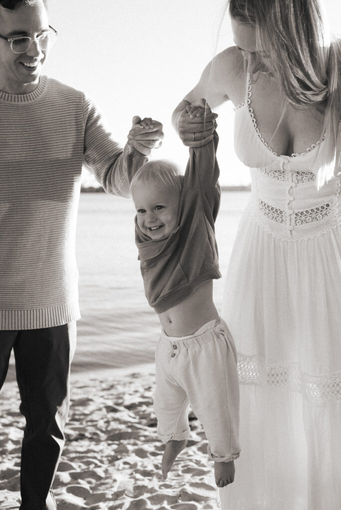 Black and white image of a family of three. The parents are holding their toddler son up by his arms while he smiles at the beach in Pacific Beach, CA