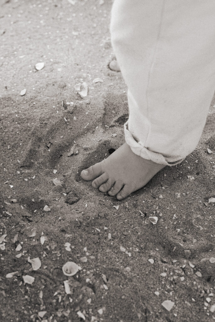 A black and white close up shot of a toddlers little feet in beach sand filled with tiny white shells