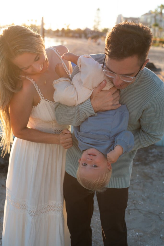 Parents hold their toddler son upside down while he giggles during golden hour at the beach in Pacific Beach, CA