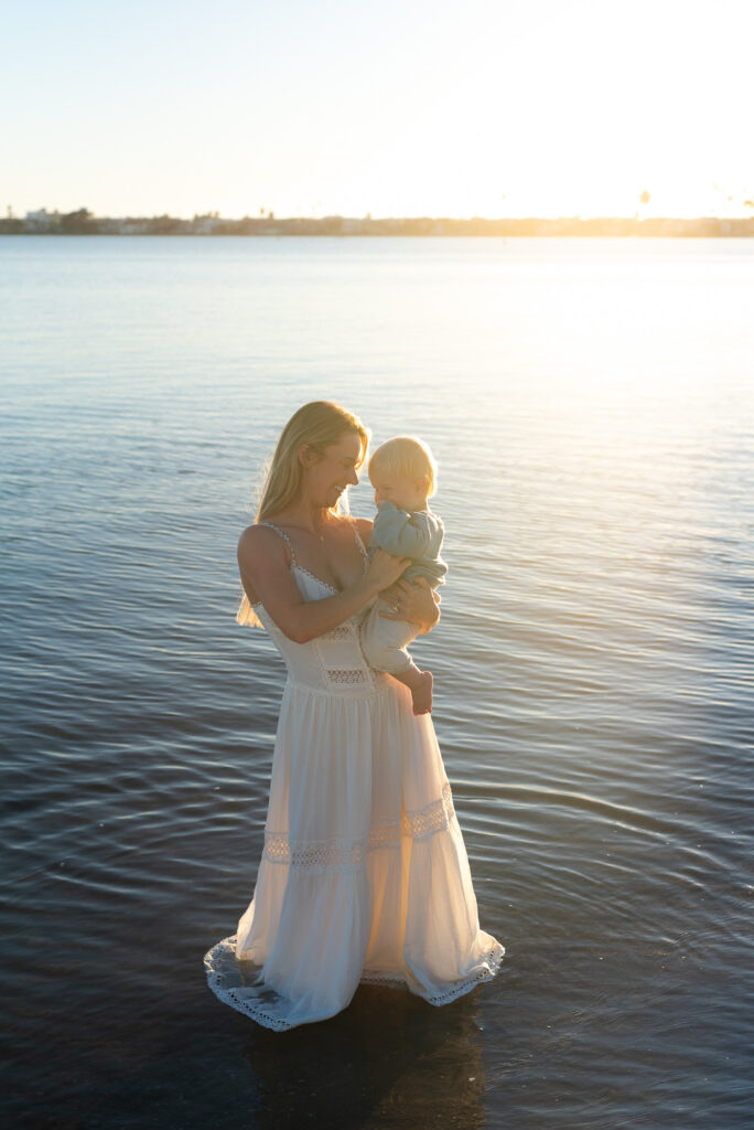 Mother stands with her toddler son during golden hour in the ocean in Pacific Beach, CA