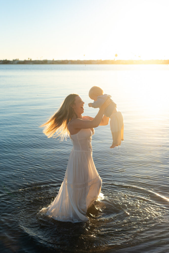 Mother is flowing white dress stands in the ocean during golden hour and spins her toddler son in Pacific Beach, CA