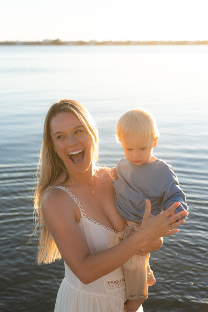 Mother holding her toddler son laughs at someone off camera with a blue ocean backdrop and golden light