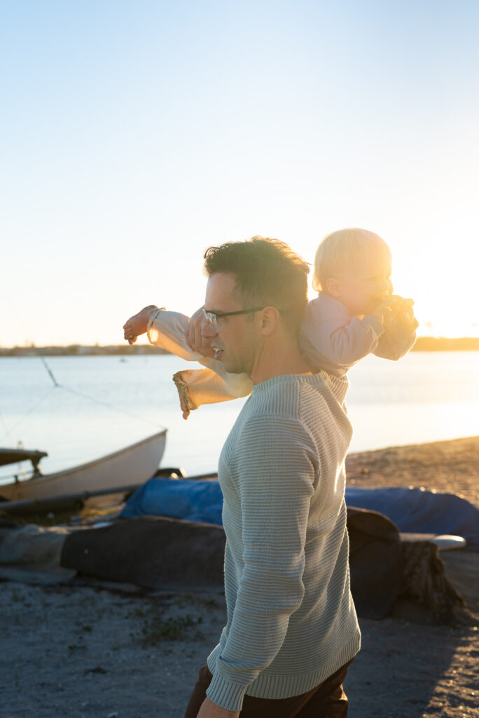 Dad carries his toddler song over his shoulder with golden light coming from behind at the beach in Pacific Beach, CA